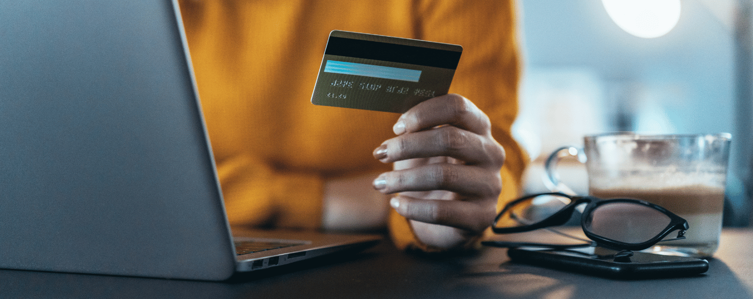 Person in Yellow Sweater Holding Credit Card Near Laptop, Glasses and Coffee Mug on Desk, Depicting Payment Gateway